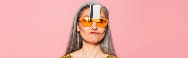 Mature asian woman in yellow sunglasses with credit card near forehead isolated on oink, banner — Stock Photo