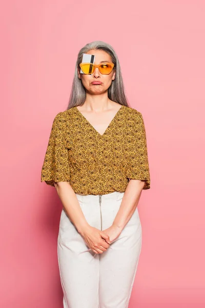 Mujer asiática madura en gafas de sol amarillas, con ojo cubierto con tarjeta de crédito mueca en rosa - foto de stock