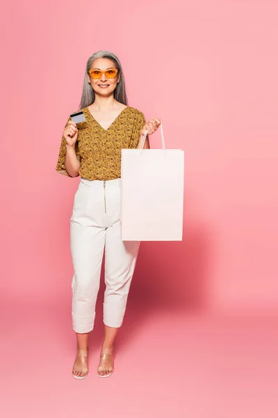 Élégante femme asiatique en pantalon blanc debout avec carte de crédit et sac à provisions sur fond rose — Photo de stock