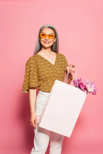 Стильная женщина средних лет с пионами в белой сумке для покупок на розовом фоне — стоковое фото