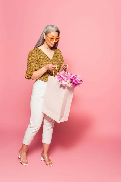 Amazed asian woman in white pants looking into shopping bag with peonies on pink background — Stock Photo