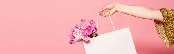 Vista cortada da mulher segurando saco de compras branco com peônias isoladas em rosa, banner — Fotografia de Stock