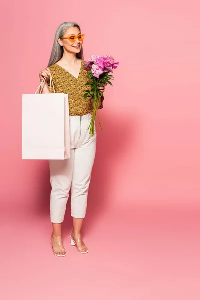 Vista completa de la mujer asiática feliz con peonías y bolsa de compras en rosa - foto de stock