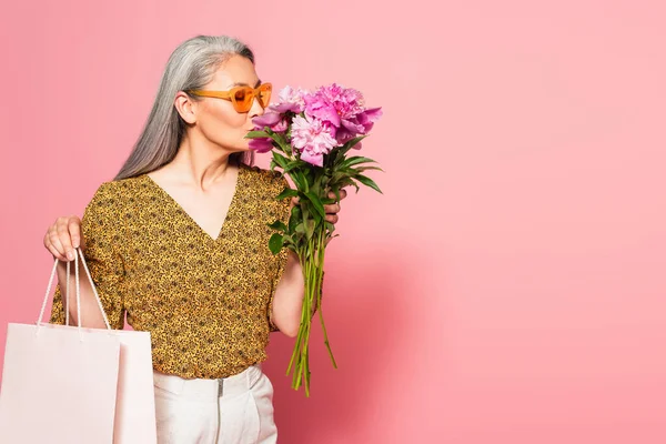 Mujer asiática madura con bolsa de compras disfrutando del aroma de peonías frescas sobre fondo rosa - foto de stock