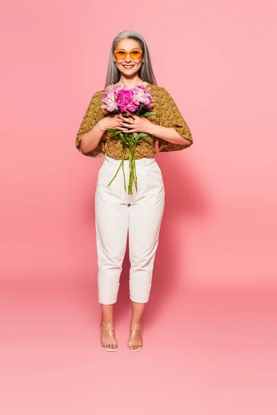 Vista completa de la mujer asiática de moda con ramo de peonías sonriendo a la cámara en rosa - foto de stock