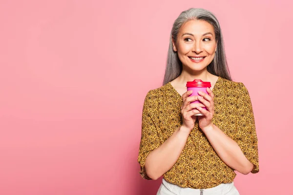 Cheerful asian woman with coffee to go looking aside on pink background — Stock Photo