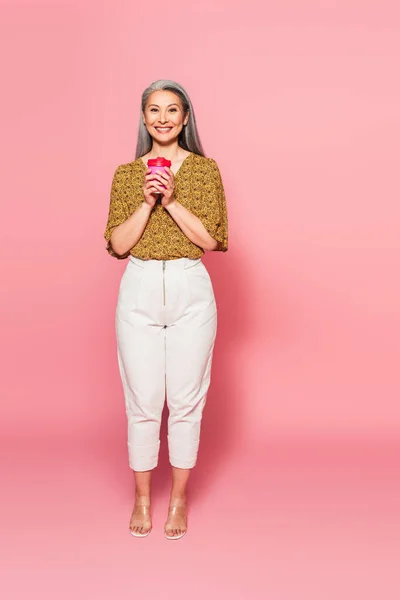 Vista completa de la mujer asiática de moda con taza de papel sobre fondo rosa - foto de stock
