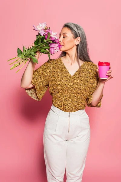 Mature asian woman with takeaway drink enjoying flavor of peonies on pink background — Stock Photo