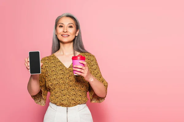 Усміхнена азіатська жінка з кавою, щоб піти, показуючи смартфон з порожнім екраном на рожевому — стокове фото