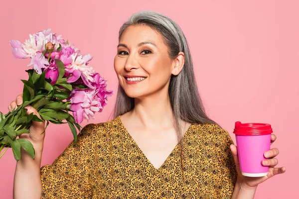 Fröhliche asiatische Frau mit Blumen und Kaffee zu gehen Blick auf Kamera isoliert auf rosa — Stockfoto
