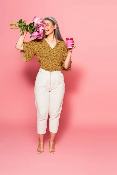 Happy asian woman with takeaway drink obscuring face with peonies on pink background — Stock Photo