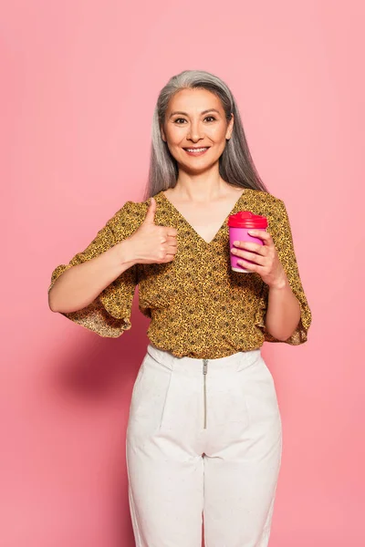 Complacido asiático mujer con café a ir mostrando pulgar arriba en rosa fondo - foto de stock
