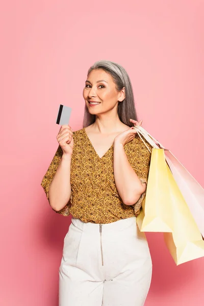 Elegante mulher asiática com sacos de compras mostrando cartão de crédito no fundo rosa — Fotografia de Stock