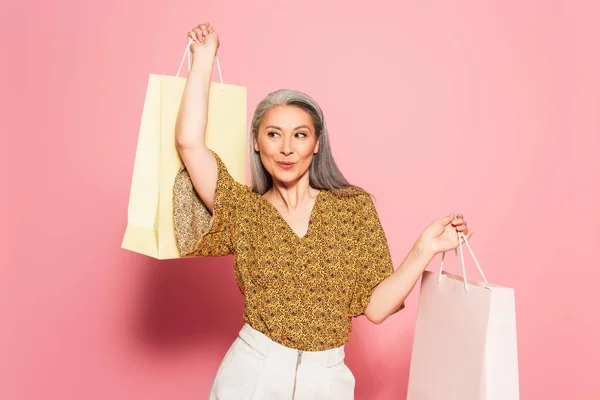 Cheerful asian woman in stylish blouse posing with shopping bags on pink background — Stock Photo