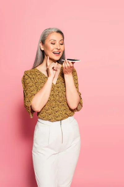 Positive asian woman in trendy clothes sending voice message on pink background — Stock Photo