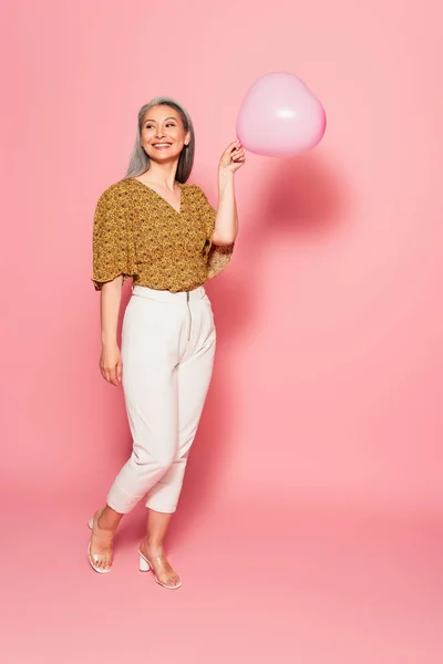 Piena lunghezza vista di sorridente donna asiatica con palloncino a forma di cuore su sfondo rosa — Foto stock