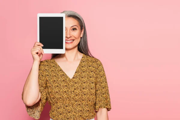 Lächelnde asiatische Frau verdeckt Gesicht mit digitalem Tablet isoliert auf rosa — Stockfoto