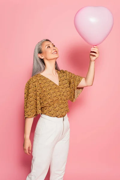 Joyful asian woman looking at heart-shaped balloon on pink background — Stock Photo