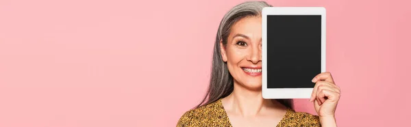 Щаслива азіатська жінка затемнює обличчя цифровим планшетом ізольовано на рожевому, банер — стокове фото