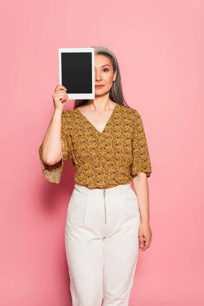 Asian woman in patterned blouse covering face with digital tablet on pink background — Stock Photo
