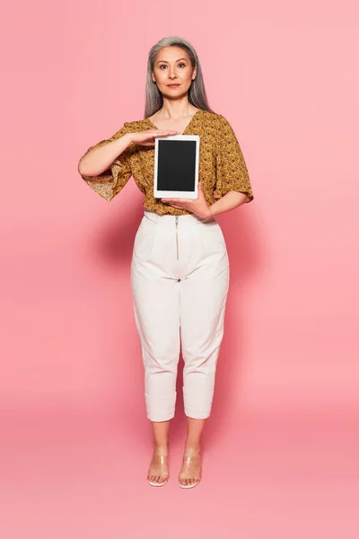 Повнометражний вид на азіатську жінку в стильному одязі, що тримає цифровий планшет з порожнім екраном на рожевому — стокове фото