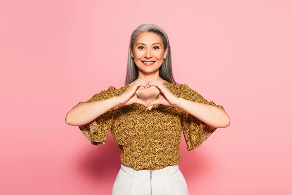 Satisfeito asiático mulher no amarelo blusa com padrão mostrando coração símbolo no rosa fundo — Fotografia de Stock