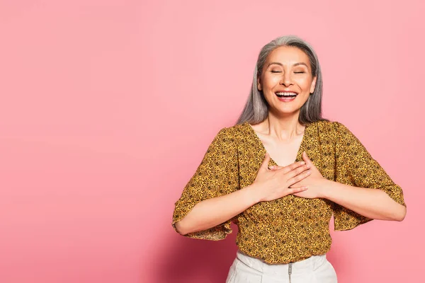 Aufgeregte asiatische Frau lacht mit geschlossenen Augen und anrührender Brust auf rosa Hintergrund — Stockfoto