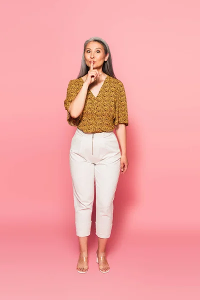 Mujer asiática en ropa de moda mirando a la cámara y mostrando el signo de silencio en rosa - foto de stock