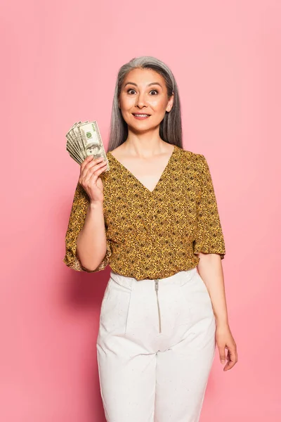 Alegre asiático mujer en de moda ropa holding dólares en rosa fondo - foto de stock