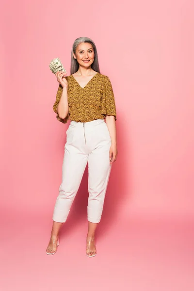 Vista a figura intera di donna asiatica in pantaloni bianchi e camicetta in piedi con banconote in dollari su rosa — Foto stock