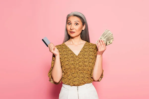 Mujer asiática divertida con tarjeta de crédito y billetes de dólar mirando a la cámara en el fondo rosa - foto de stock