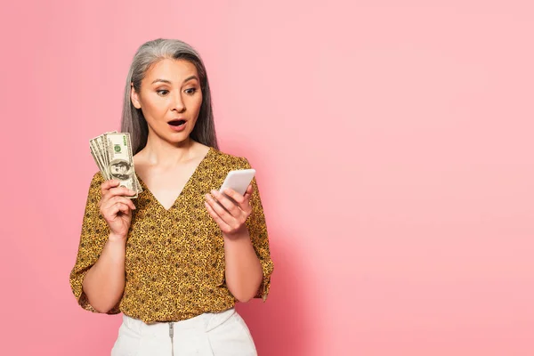 Sorprendida mujer asiática con billetes de dólar mirando el teléfono inteligente sobre fondo rosa - foto de stock