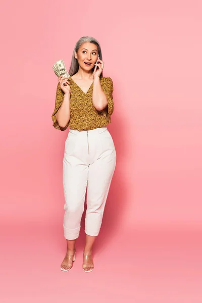 Full length view of amazed asian woman with money talking on smartphone on pink — Stock Photo