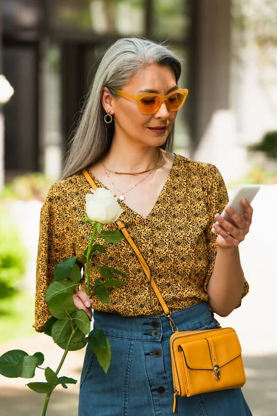 Middle aged asian woman in trendy clothes holding white rose while messaging on smartphone outdoors — Stock Photo