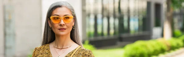 Зріла азіатська жінка в жовтих сонцезахисних окулярах дивиться далеко на міську вулицю, банер — стокове фото