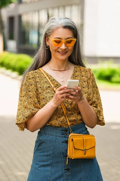 Веселая азиатка в солнечных очках и стильной одежде, печатающей на мобильном телефоне на открытом воздухе — стоковое фото