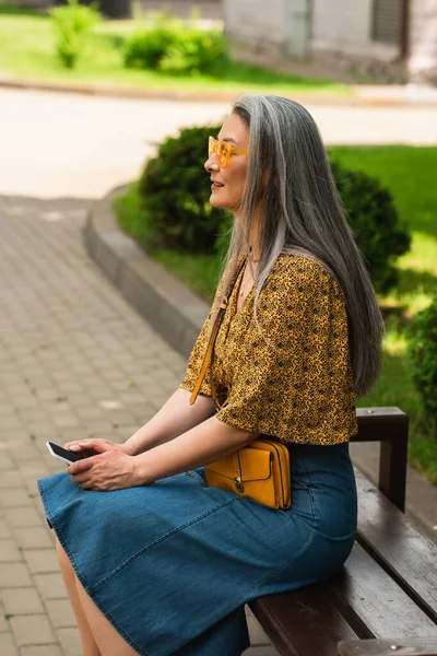 Middle aged asian woman in trendy clothes sitting on city bench with smartphone — Stock Photo