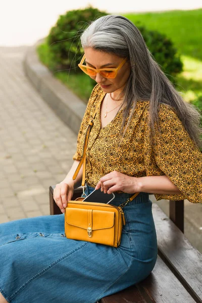 Trendy asian woman with crossbody bag and smartphone sitting on bench in city — Stock Photo