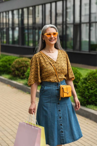 Sonriendo mujer asiática en ropa de moda y gafas de sol caminando con bolsas de compras al aire libre - foto de stock