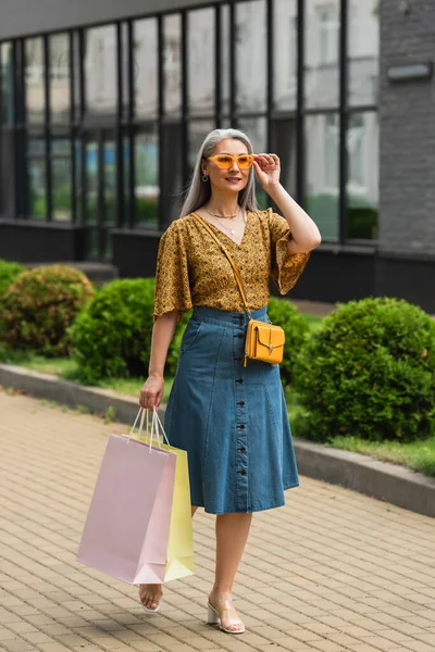 Fashionable asian woman adjusting sunglasses while walking with shopping bags along city street — Stock Photo