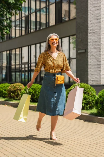 Радісна азіатська жінка в джинсовій спідниці і візерункова блузка, що йде з сумками на вулиці — стокове фото