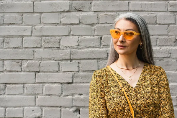 Reife asiatische Frau mit stylischer Sonnenbrille, die in der Nähe einer Ziegelmauer in der Stadt wegschaut — Stockfoto