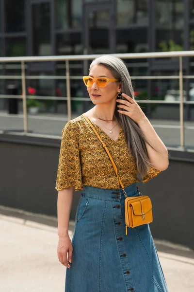 Mujer asiática de mediana edad en ropa de moda ajustando el pelo gris mientras mira al aire libre - foto de stock