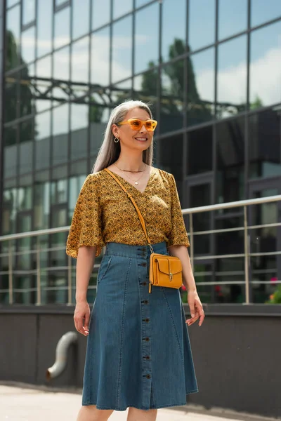 Alegre asiático mujer en moda blusa y denim falda mirando lejos al aire libre - foto de stock