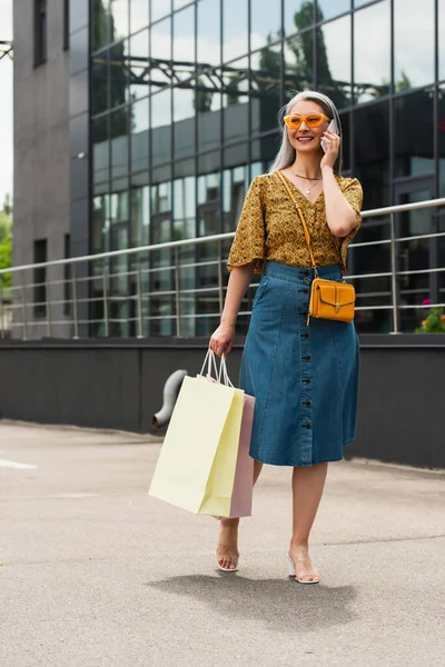 Mature asian woman in trendy blouse and denim skirt talking on cellphone while holding shopping bags — Stock Photo
