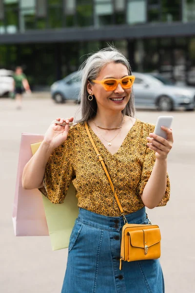 Cheerful asian woman in patterned blouse and sunglasses using cellphone while standing with shopping bags — Stock Photo