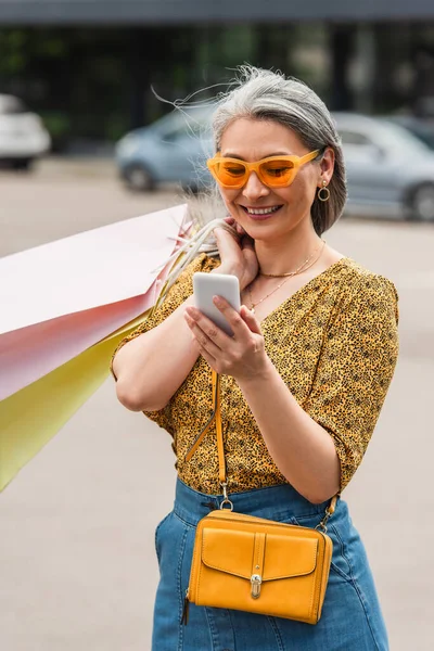 Trendige asiatische Frau mit Einkaufstaschen, die auf der Straße auf ihr Handy schaut — Stockfoto