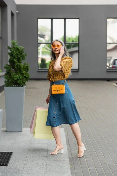 Stilvolle Asiatin mit Einkaufstüten telefoniert in der Nähe von Gebäude — Stockfoto