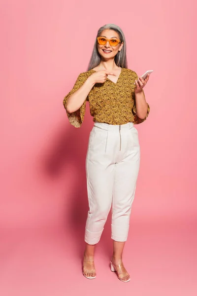 Mature asian woman in stylish clothes and sunglasses pointing at smartphone on pink background — Stock Photo