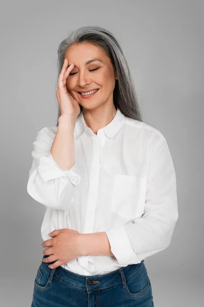 Mature asian woman in white shirt touching face while laughing with closed eyes isolated on grey — Stock Photo